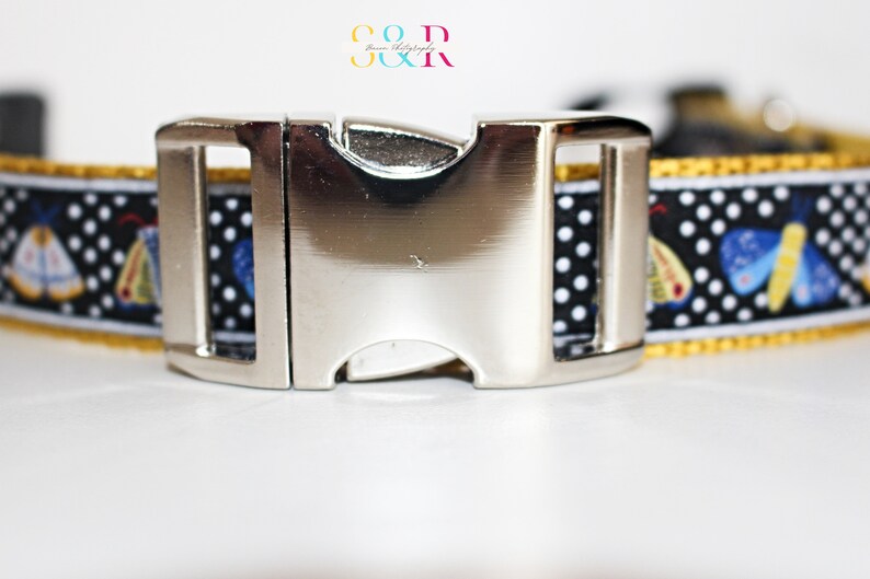 May include: A black and white dog collar with a silver buckle. The collar has a white polka dot pattern and a yellow trim. The collar features a repeating pattern of blue and yellow butterflies.