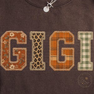 May include: A dark brown sweatshirt featuring the word GIGI in large, fabric-appliqued letters. Each letter has a distinct pattern: floral, leopard print, orange plaid, and green gingham. The letters are outlined with a decorative zigzag stitch.