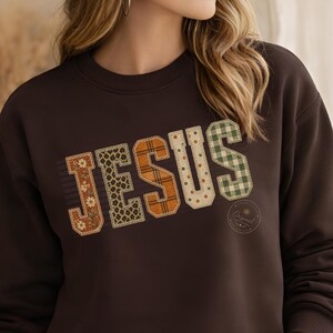 May include: A dark brown crewneck sweatshirt featuring the word JESUS in large, block-style letters. Each letter has a unique pattern, including floral, leopard print, plaid, polka dot, and green gingham, all outlined with a decorative stitch.
