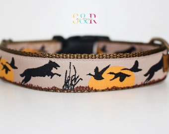 May include: A tan dog collar with a black silhouette of a dog running and ducks flying in front of a setting sun. The collar has a brown trim and a black buckle.