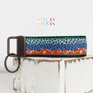 May include: A key fob with a brown, blue, and orange fish scale pattern. The key fob has a dark metal clasp and key ring. The key fob is on a white, textured surface. The text "S & R" and "Baron Photography" is in the background.