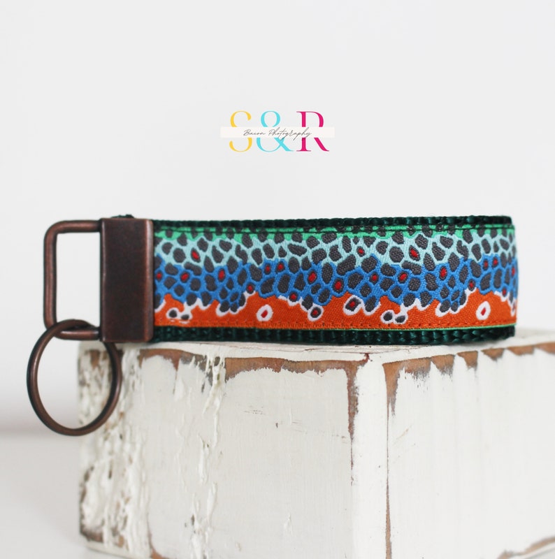 May include: A key fob with a brown, blue, and orange fish scale pattern. The key fob has a dark metal clasp and key ring. The key fob is on a white, textured surface. The text "S & R" and "Baron Photography" is in the background.