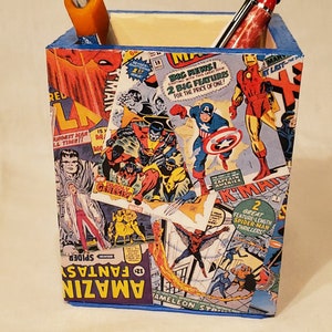 Puede incluir: Un porta lápices de madera con un borde azul, cubierto de un collage de portadas de cómics vintage con personajes de Marvel como Spider-Man, Capitán América y Iron Man. El texto en las portadas incluye "Amazing Fantasy", "Captain America", "Iron Man" y "Spider-Man".