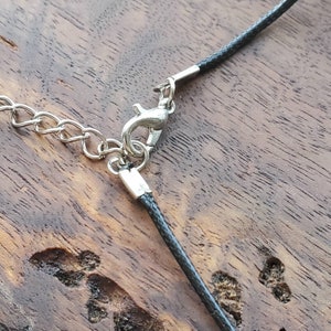 May include: Close-up of a black cord necklace with a silver clasp. The cord is lying on a wooden surface.