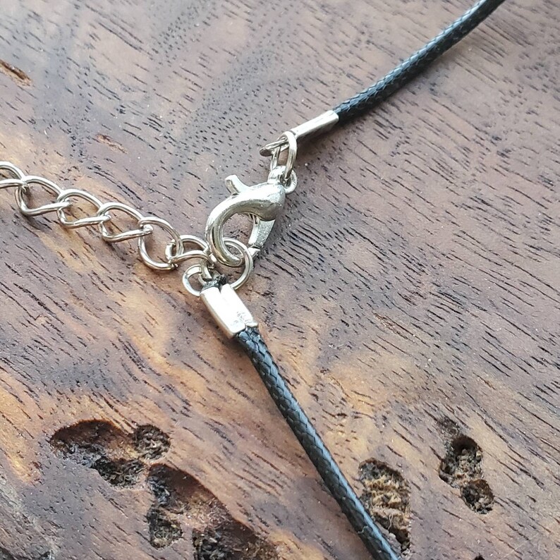 May include: Close-up of a black cord necklace with a silver clasp. The cord is lying on a wooden surface.