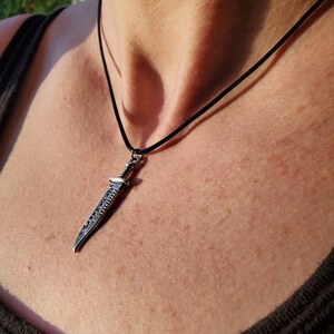 May include: A silver sword pendant necklace on a black cord. The sword has a detailed design and is worn on a person's chest.