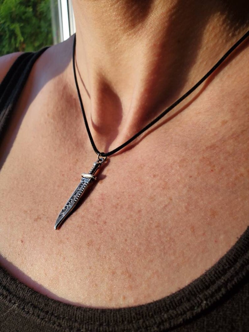 May include: A silver sword pendant necklace on a black cord. The sword has a detailed design and is worn on a person's chest.