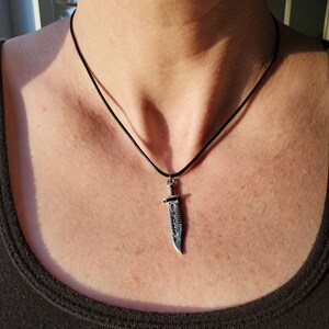 May include: A black cord necklace with a silver knife pendant. The knife has a detailed design and a sharp point.