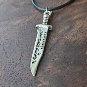 May include: A silver-toned pendant necklace featuring a detailed knife design with an inscription on the blade. The necklace is on a black cord.