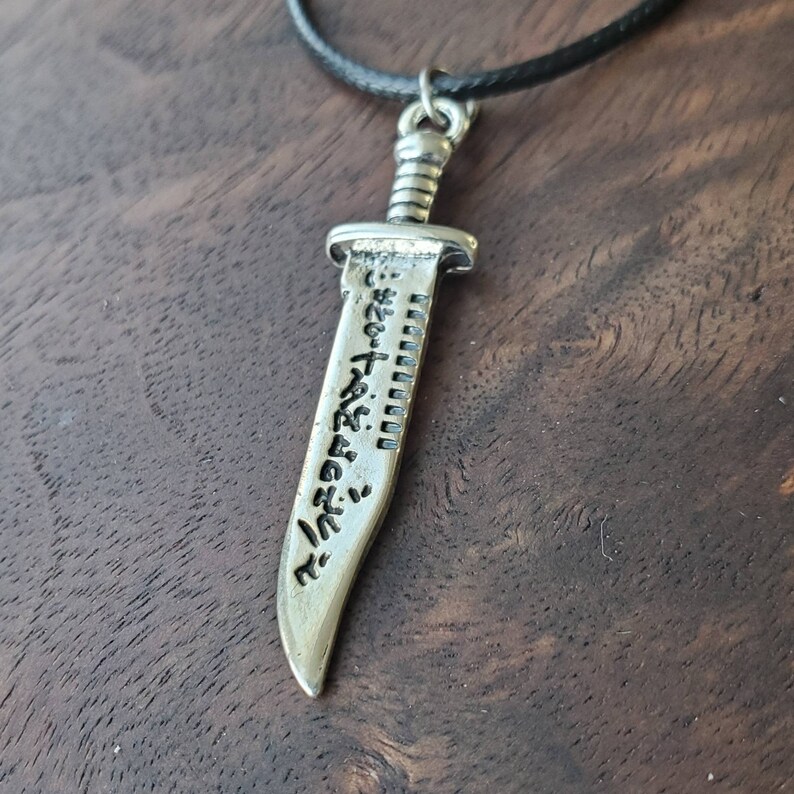 May include: A silver-toned pendant necklace featuring a detailed knife design with an inscription on the blade. The necklace is on a black cord.