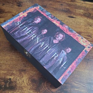 May include: Black wooden box featuring the Supernatural logo and an image of the main characters from the TV show. The logo is in white text with a fiery orange background.