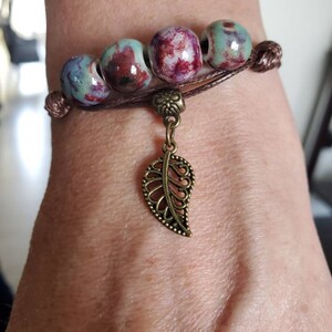 Op de afbeelding: Een bruin leren armband met een bedel in de vorm van een blad. De armband heeft vijf keramische kralen in een mix van rood, wit en blauw.