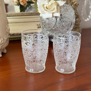 May include: Two clear, vintage-style drinking glasses with an ornate, swirling pattern. The glasses are cylindrical with a slightly flared base, and sit on a wooden surface. The background includes a decorative vase and mirror.