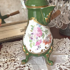 May include: A green porcelain chocolate pot with a lid and a wooden handle. The body features a white central panel decorated with a colourful floral bouquet of pink roses and purple flowers, accented by gold scrollwork and three ornate feet.