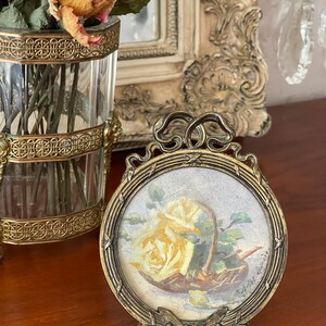 Può includere: Una cornice rotonda vintage color ottone con un'immagine dipinta di una rosa gialla in un cesto. La cornice ha un design decorativo e un supporto. Un vaso di vetro con rose secche è sullo sfondo.