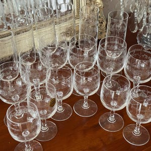 Peut inclure: Une collection de verres à vin et de grands verres à boire en verre clair gravé. Les verres à vin ont un bol arrondi et une tige. Les grands verres sont cylindriques. Les verres sont disposés sur une surface en bois foncé.