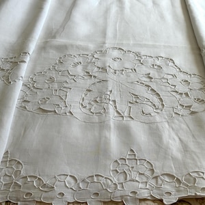 May include: White linen tablecloth with intricate cutwork floral and leaf designs. The cutwork is along the edges and in a large central panel. The fabric is folded, showing the texture and detail of the embroidery.