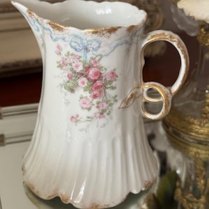 Op de afbeelding: Witte porseleinen creamer met een bloemmotief van roze rozen en een blauw lint. De creamer heeft een goudkleurige handgreep en rand, en een gegroefde body. Een decoratief stuk voor het serveren van room of melk.