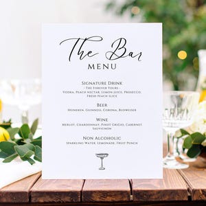 Może przedstawiać: Białe menu barowe z elegancką kaligrafią, zawierające drinki firmowe, piwo, wino i opcje bezalkoholowe. Menu znajduje się na ciemnym drewnianym stole, z kieliszkami, kwiatami i zielenią w tle.