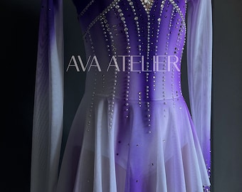 Puede incluir: Un vestido de patinaje artístico morado y blanco con mangas largas y una falda blanca. El vestido está decorado con pedrería y el texto "AVA ATELIER" está impreso en la parte delantera.