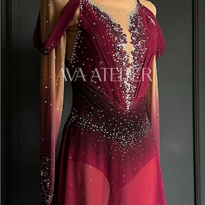 May include: A burgundy figure skating dress with a gradient effect, adorned with sparkling embellishments. The dress features long sleeves, a sheer neckline, and the text "AVA ATELIER". The design includes a skirt and shoulder drapes.