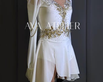 May include: White figure skating dress with long sheer sleeves and a split skirt. The bodice and waist are embellished with gold and clear rhinestones. The text "AVA ATELIER" is printed on the dress.