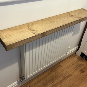 May include: A wooden shelf mounted above a white radiator. The shelf is stained a light brown colour and has a natural wood grain.