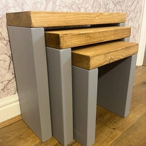 May include: Set of three nesting tables with a grey painted base and natural wood tops. The tables are stacked on top of each other.