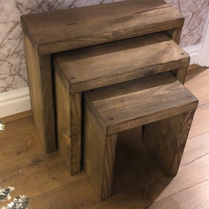 May include: A set of three wooden nesting tables in a dark brown finish. The tables are made of solid wood and have a rustic look.