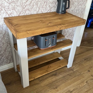 May include: A white and brown wooden kitchen island with two shelves. A toaster and a kettle are on the top shelf. The island is in a room with light brown wood flooring.
