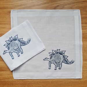 Könnte beinhalten: Zwei quadratische weiße Servietten mit einer dunkelblauen Strichzeichnung eines Stegosaurus-Dinosauriers. Die größere Serviette liegt flach, während die kleinere Serviette gefaltet ist und teilweise auf der größeren liegt, beide mit dem Motiv.
