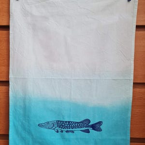 Può includere: Un canovaccio da cucina bianco e blu sfumato con un'illustrazione di pesce blu scuro. Il disegno del pesce è stampato nella metà inferiore del canovaccio. Il canovaccio è appeso con una molletta blu.