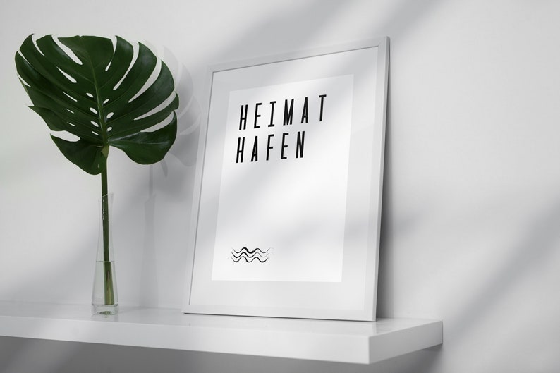 K&ouml;nnte beinhalten: Ein wei&szlig;er gerahmter Druck mit dem Text "HEIMAT  HAFEN" und einem Wellenlinien-Design. Der Druck befindet sich auf einem wei&szlig;en Regal mit einer gr&uuml;nen Pflanze in einer durchsichtigen Glasvase.