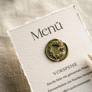 May include: A menu card with the word "Men&uuml;" at the top, featuring a decorative wax seal with a wreath design. The menu lists "VORSPEISE" and other German food items. The card has a torn edge and is on a textured surface.