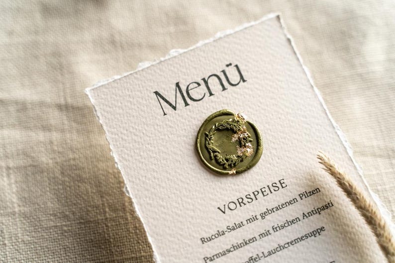 May include: A menu card with the word "Men&uuml;" at the top, featuring a decorative wax seal with a wreath design. The menu lists "VORSPEISE" and other German food items. The card has a torn edge and is on a textured surface.