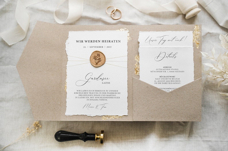 Op de afbeelding: Een huwelijksuitnodiging met een rustieke bruine papieren envelop en een witte kaart met gouden accenten. De kaart heeft de tekst "Wir werden heiraten" (We gaan trouwen) en de datum "18. September 2022". De kaart is verzegeld met een waszegel met een bloemmotief.