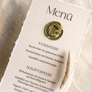 May include: A menu card with a torn edge, featuring the word "Men&uuml;" at the top. The card has a gold seal and lists appetizers, main courses, and desserts. A sprig of wheat is placed on the right side.
