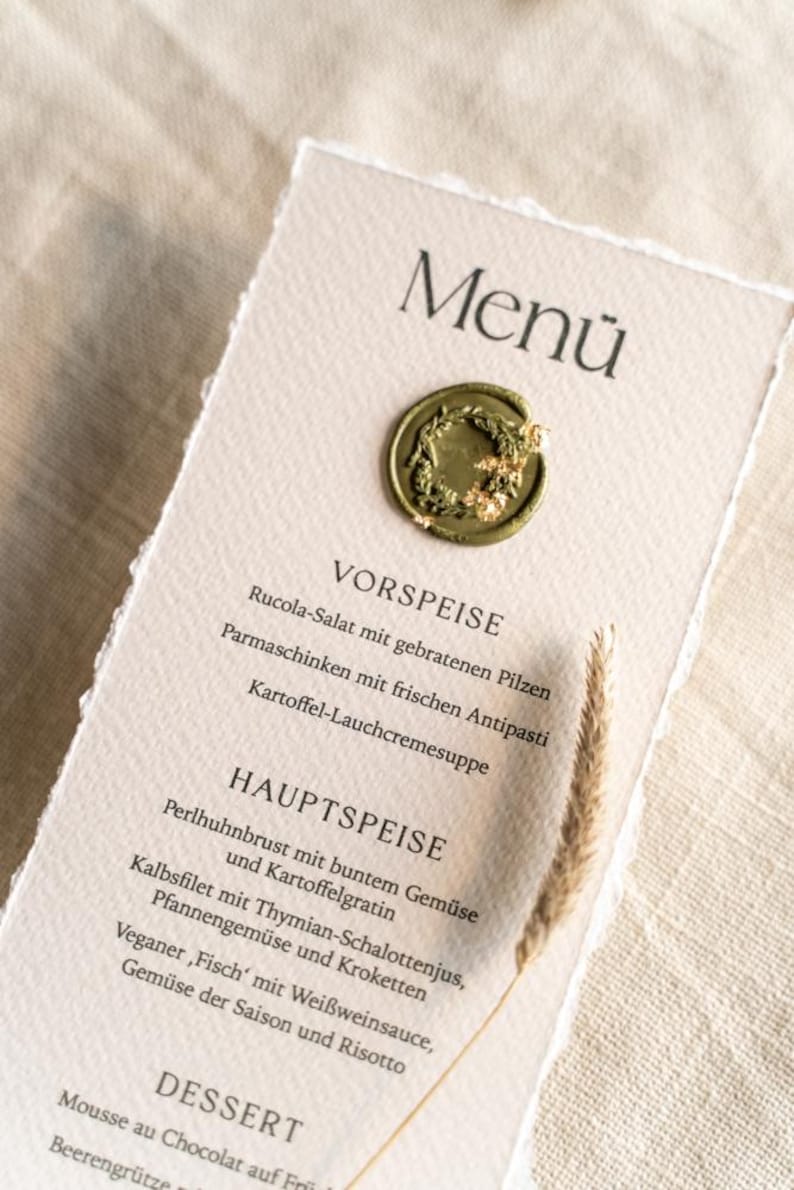 May include: A menu card with a torn edge, featuring the word "Men&uuml;" at the top. The card has a gold seal and lists appetizers, main courses, and desserts. A sprig of wheat is placed on the right side.