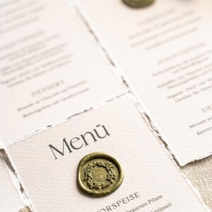 May include: Close-up of elegant menu cards with a vintage aesthetic. Each card features the word "Menu" and is sealed with a gold wax stamp. The menu items are printed in a classic font, suggesting a fine dining experience.
