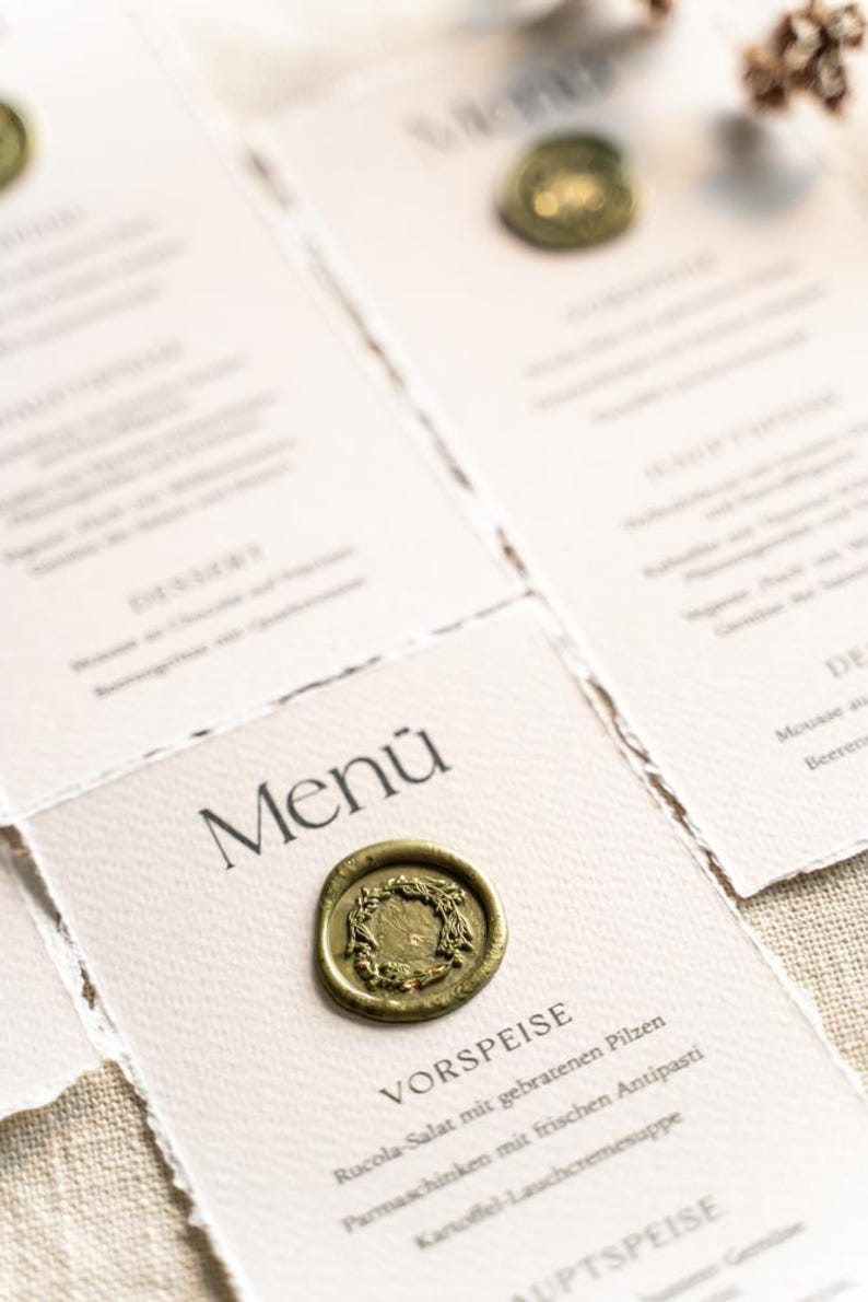 May include: Close-up of elegant menu cards with a vintage aesthetic. Each card features the word "Menu" and is sealed with a gold wax stamp. The menu items are printed in a classic font, suggesting a fine dining experience.