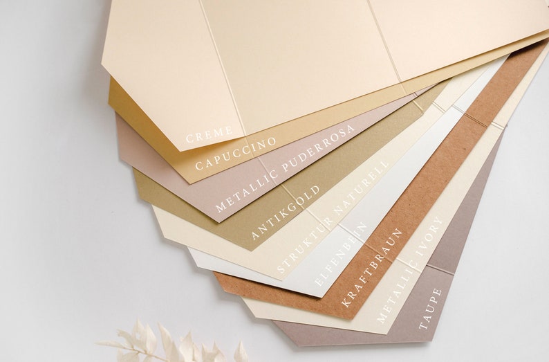 Op de afbeelding: Een verzameling papierstalen in verschillende tinten beige, bruin en roze. De papierstalen zijn gelabeld met namen zoals "Creme", "Cappuccino", "Metallic Puderrosa", "Antikgold", "Struktur Naturell", "Elfenbeen", "Kraftbraun", "Metallic Ivory" en "Taupe".