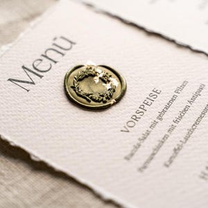 May include: A menu card with the word "Men&uacute;" at the top, sealed with a gold wax seal. The menu features German text, including "VORSPEISE" and "HAUPTSPEISE", indicating appetizers and main courses. The card is on a textured, off-white paper.