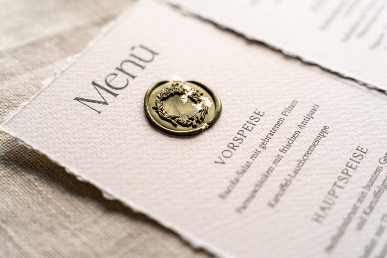 May include: A menu card with the word "Men&uacute;" at the top, sealed with a gold wax seal. The menu features German text, including "VORSPEISE" and "HAUPTSPEISE", indicating appetizers and main courses. The card is on a textured, off-white paper.