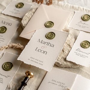 May include: Elegant wedding stationery set featuring cream-colored cards and an envelope, sealed with gold wax stamps. The cards include menu, save-the-date, and reserved seating details. The text "Martha & Leon" is visible on one card.