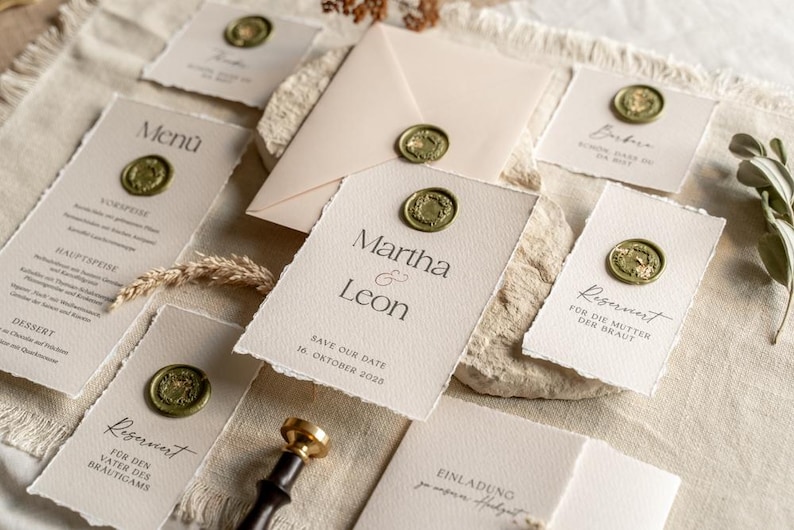 May include: Elegant wedding stationery set featuring cream-colored cards and an envelope, sealed with gold wax stamps. The cards include menu, save-the-date, and reserved seating details. The text "Martha & Leon" is visible on one card.