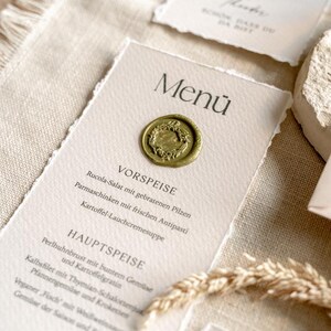 May include: Elegant wedding menu cards with a rustic aesthetic. The cards feature a torn-edge design, adorned with a gold wax seal and printed menu details. The cards are placed on a textured beige linen tablecloth.