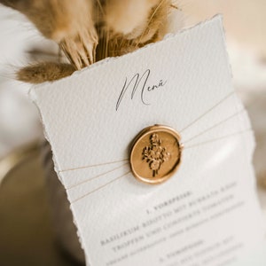 Op de afbeelding: Een wit papieren menu met een gouden waszegel en het woord "Menu" in cursief. Het menu is vastgebonden met touw en heeft een rustieke, handgemaakte uitstraling.