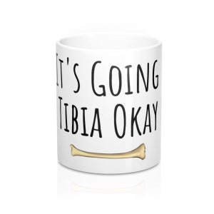 May include: White ceramic mug with black text that reads "It's going tibia okay" and a small gold bone graphic.