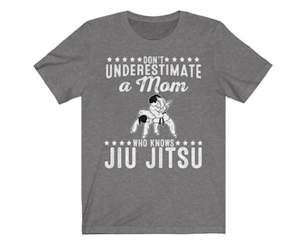 Può includere: Maglietta grigia con una grafica bianca che dice "Non sottovalutare una mamma che conosce il Jiu Jitsu". La grafica include anche un'illustrazione in bianco e nero di due persone che praticano Jiu Jitsu.