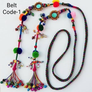 Può includere: Una cintura colorata e fatta a mano con dettagli intricati. La cintura presenta un cordoncino intrecciato nei toni del marrone, verde e viola. È adornata con pompon, perline, conchiglie ed elementi decorativi. Il testo "Belt Code-1" è visibile.